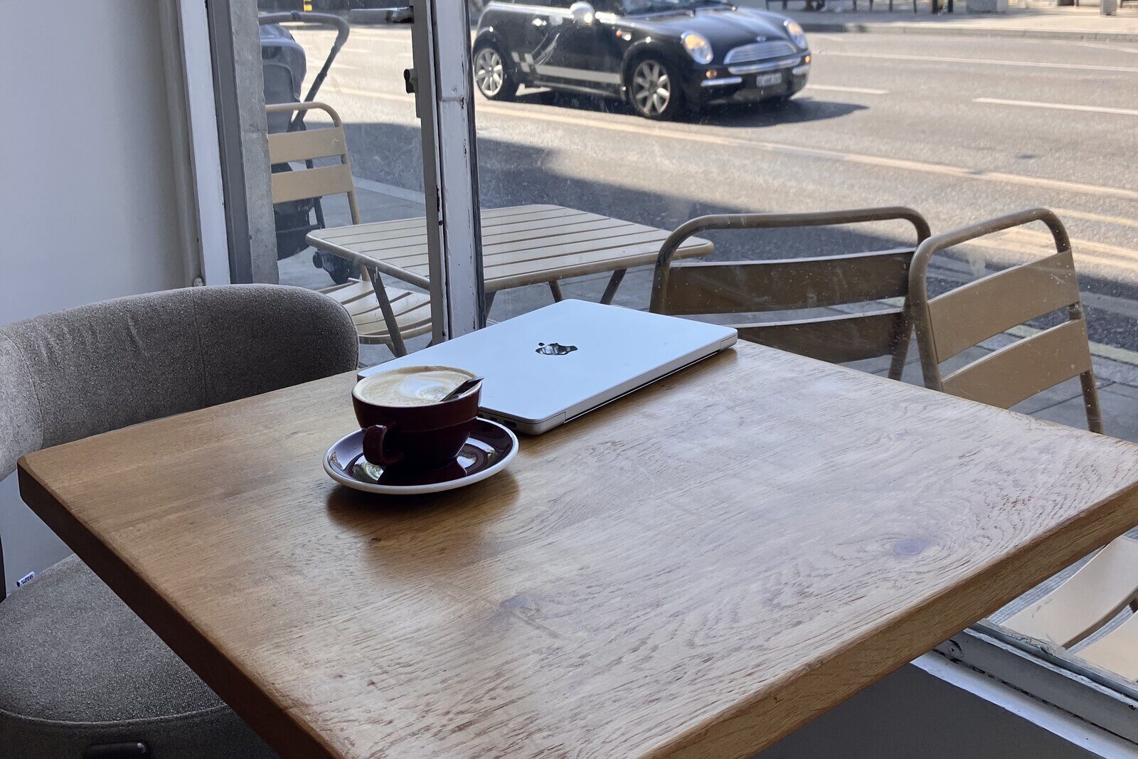 A café table by a window: a cup of coffee, a closed laptop, and a street beyond the glass.
