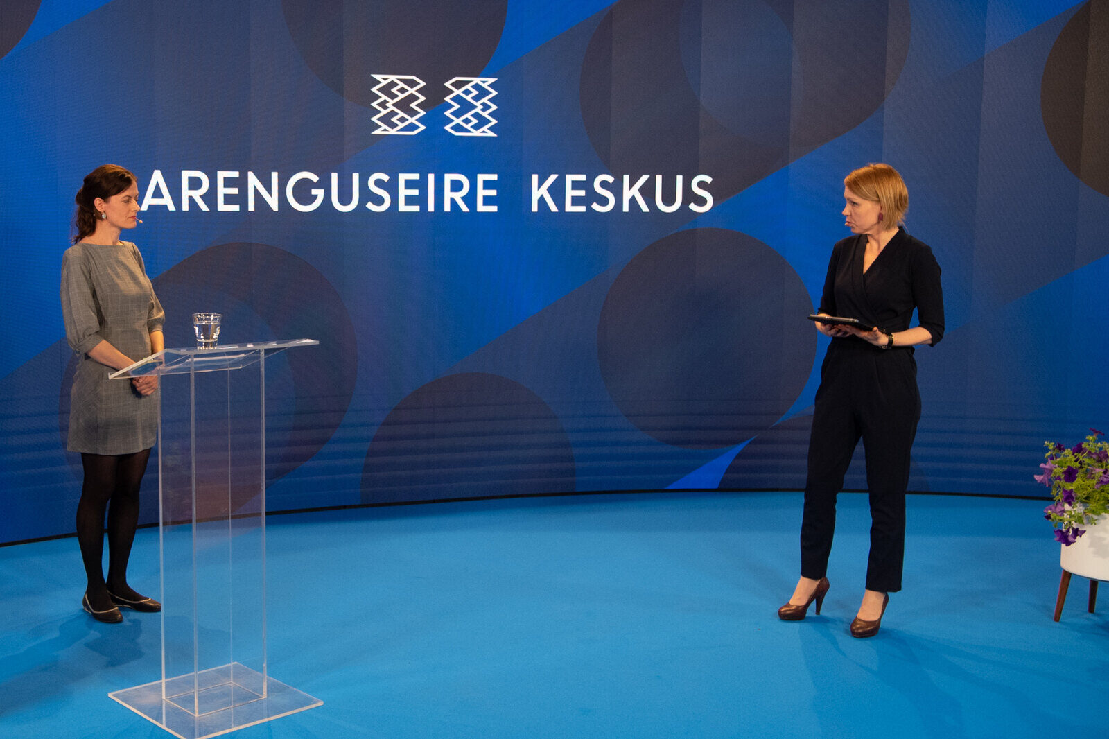 A television studio in blue: a woman at a clear lectern, mid-sentence, facing an interviewer standing a few metres away.
