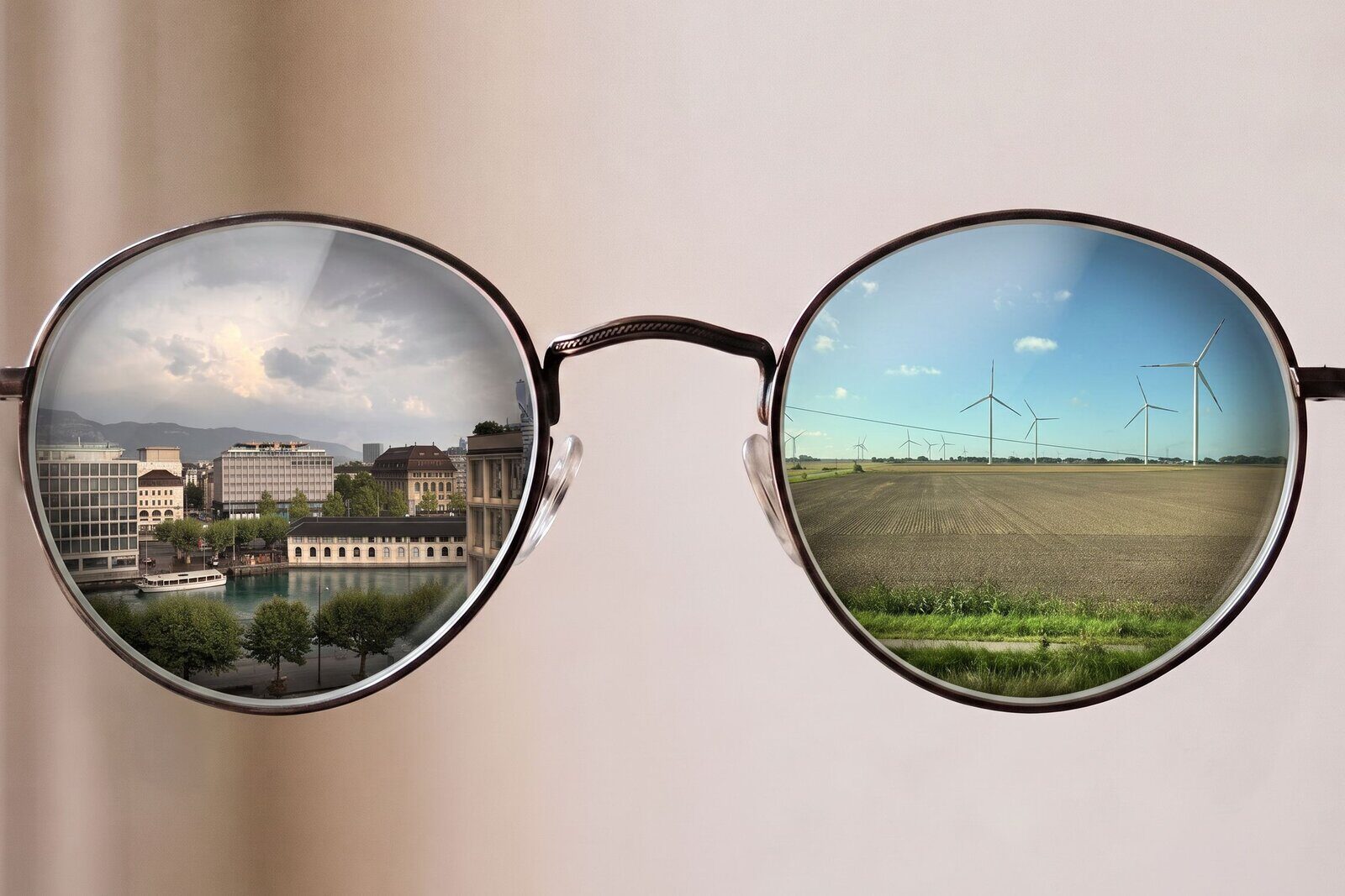 A pair of round spectacles against a plain wall; the left lens shows Geneva from above with the Rhône and the quays, the right shows a wind farm over open farmland.